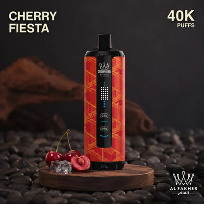 A Crown Bar disposable vape device in Cherry Fiеста flavor, featuring a red and orange geometric pattern with a black central control panel. The vape is displayed on a wooden block surrounded by fresh cherries and ice cubes, set against a dark background of pebbles and wood logs.