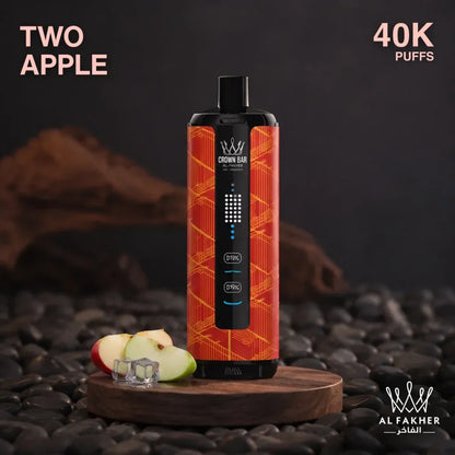 Al fakher Crown Bar disposable vape device in the 'Two Apple' flavor, featuring a black body with a vibrant orange and red geometric patterned outer shell. The vape is displayed on a wooden stand over dark pebbles, with sliced apple pieces and ice cubes nearby.